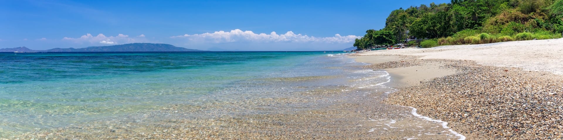 A pebbly beach with clear turquoise waters, a sandy shore, and a forested hill in the background.