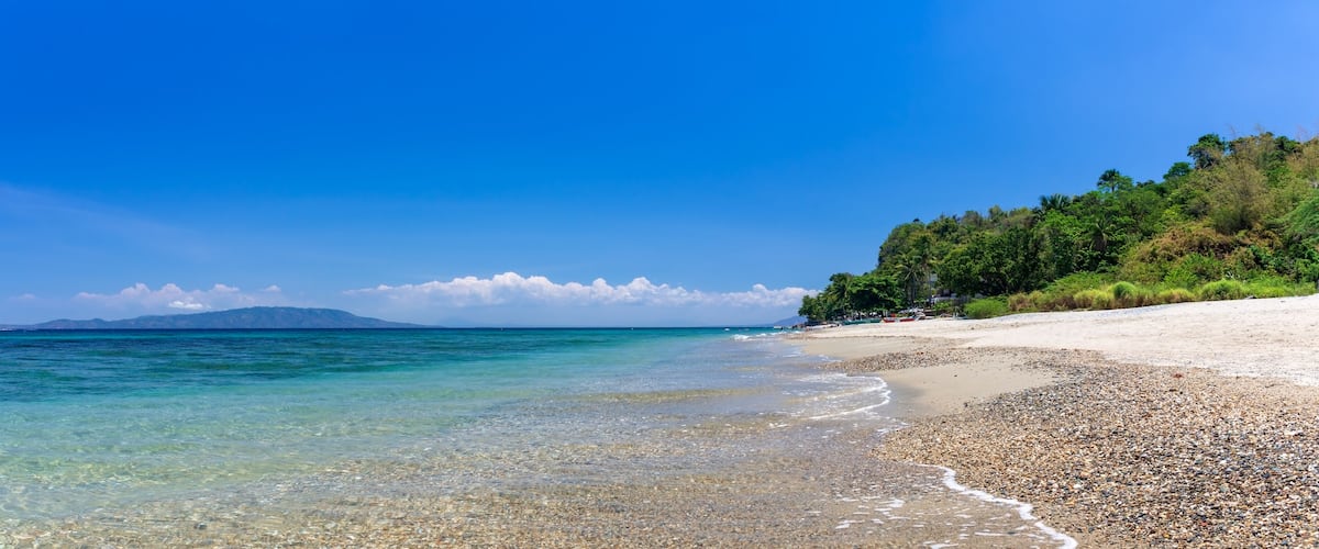 A pebbly beach with clear turquoise waters, a sandy shore, and a forested hill in the background.