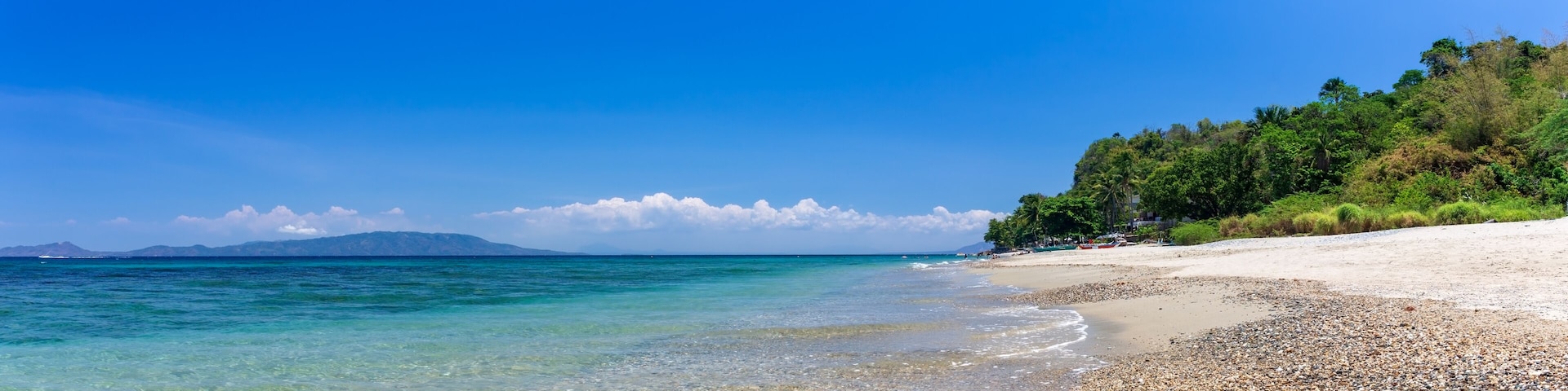 A pebbly beach with clear turquoise waters, a sandy shore, and a forested hill in the background.