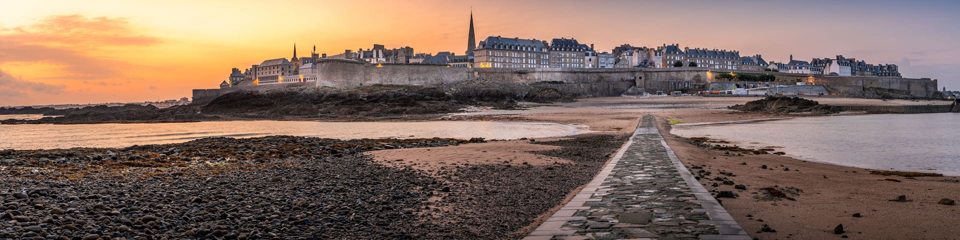 A coastal cityscape at sunset with a prominent castle and a stone pathway leading to the beach.