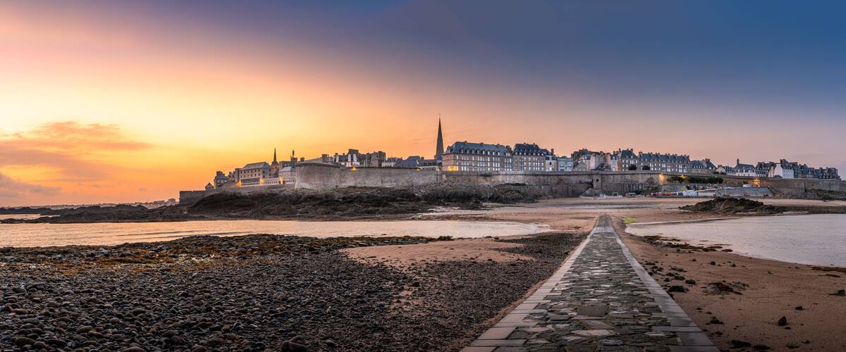 A coastal cityscape at sunset with a prominent castle and a stone pathway leading to the beach.