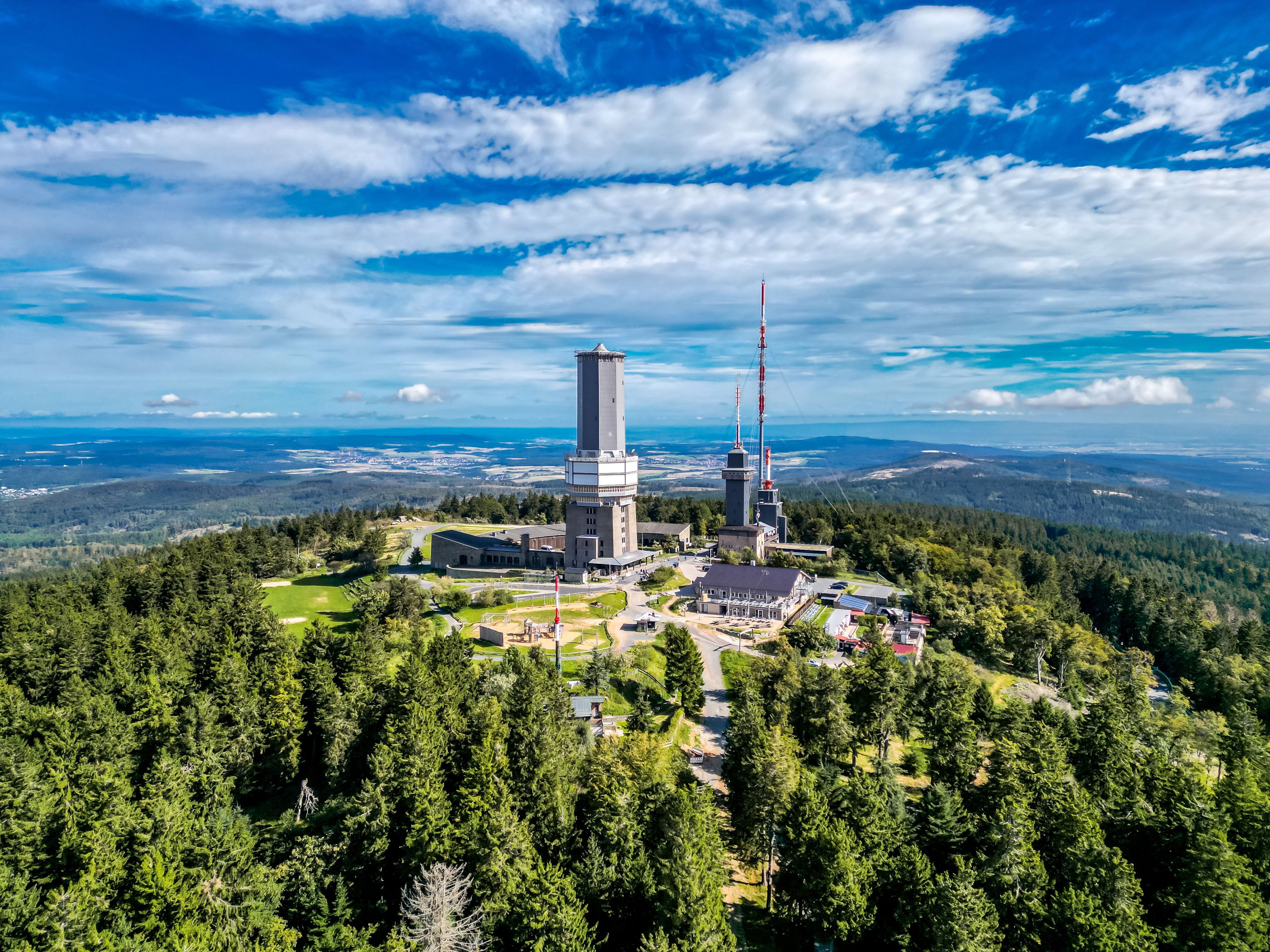 A tower with a tall antenna, surrounded by a forested area and a clear sky.