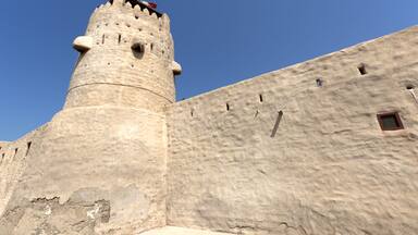 A large stone tower with a flag on top, part of a fortress with a crenellated wall and small windows.