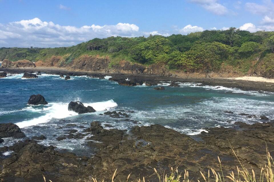 A coastal landscape with rocky shores, waves crashing against the rocks, and a backdrop of green hills under a clear sky.