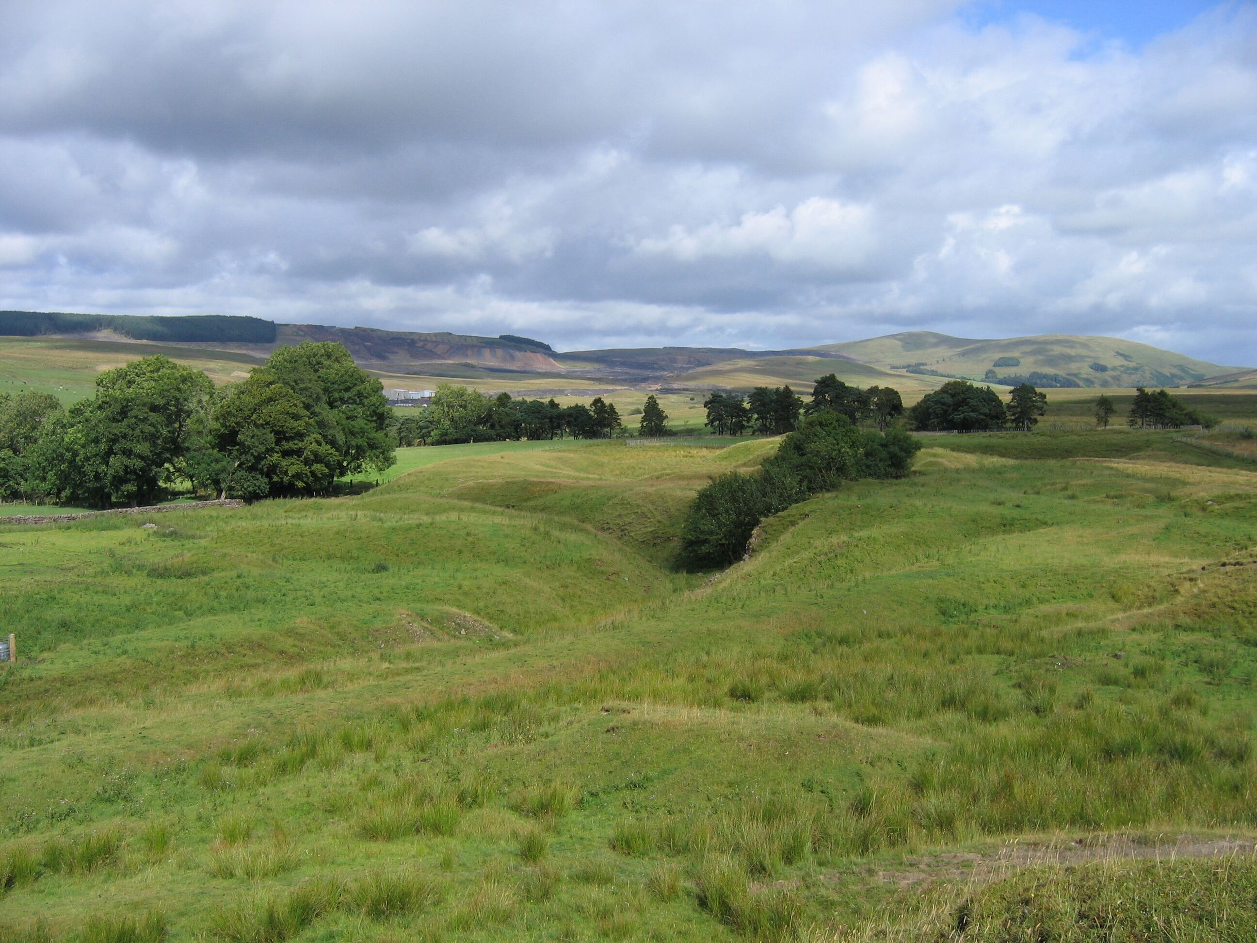 Ashieburn Quarry One of the waste mounds surrounding Ashieburn Quarry (note the older spelling of todays Ashaw Burn) gives an overview of the railway approach to the site. The branch siding from Bairds Muirkirk to Glenbuck line comes up through the trees on the left, crosses a farm track (marked by the stone wall) and curves through a right-angle past some ruined limekilns and out of the picture on the left. The level upper siding from a reversing neck, probably provided to allow coal brought by rail to be tipped into the top of the kilns, comes in alongside and heads diagonally into centre right of the picture. Short barrow runs from the siding to the tops of the kilns can be seen, the kilns being below the clump of trees in the centre of the picture. The opencast site that destroyed Glenbuck village is in the background; the nearer parts have already been restored, though old railway alignments and all other above-ground structures have been lost for ever.