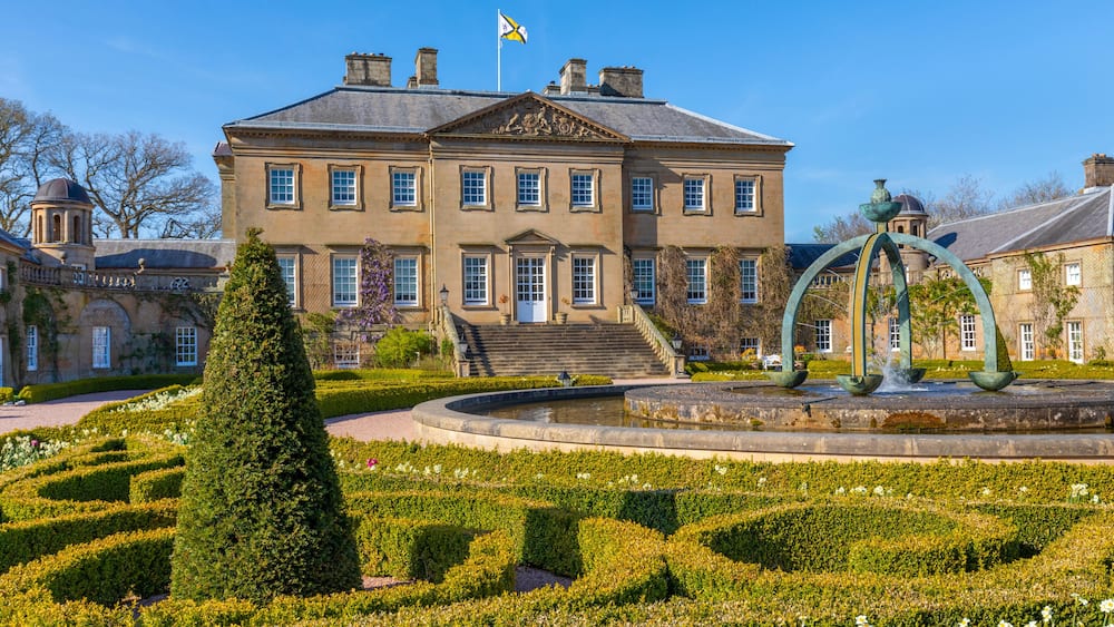 A large stately building with a flag, a formal garden with a fountain, and a neatly trimmed hedge.