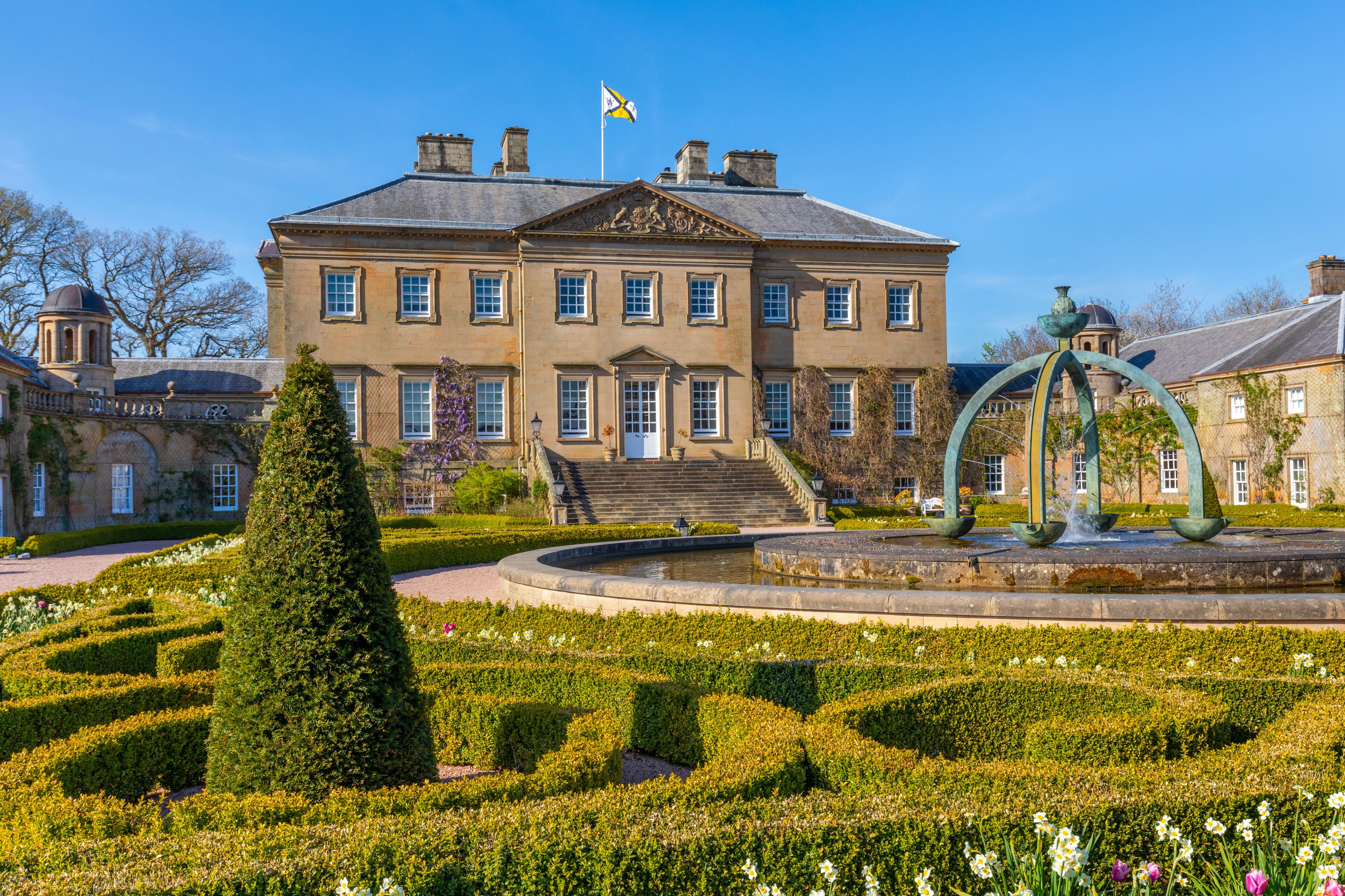 A large stately building with a flag, a formal garden with a fountain, and a neatly trimmed hedge.