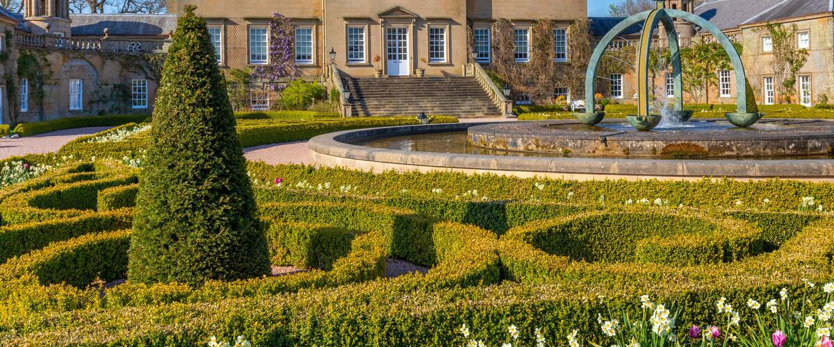 A large stately building with a flag, a formal garden with a fountain, and a neatly trimmed hedge.