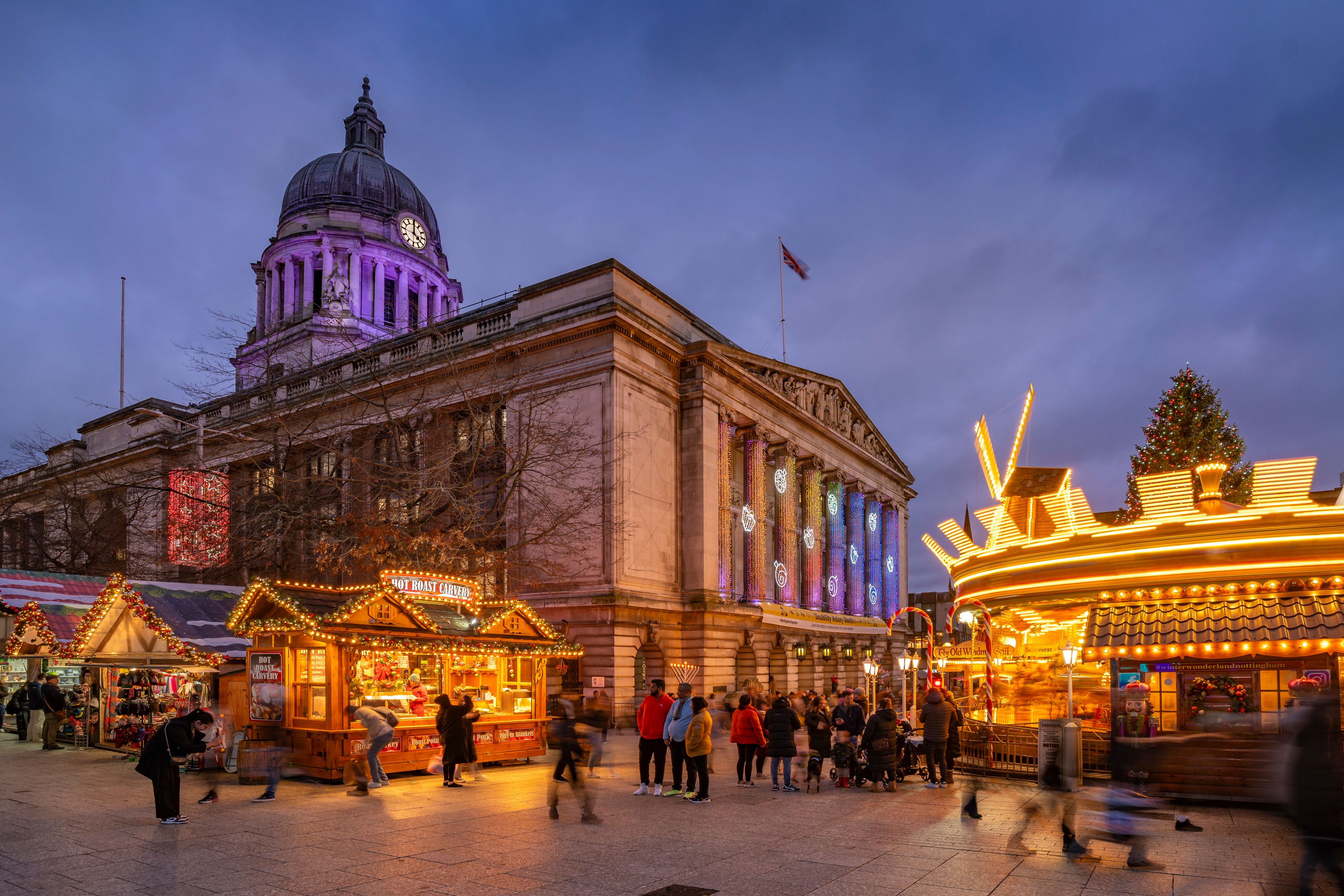 A festive market with illuminated stalls, a historic building with a dome, and a Christmas tree.