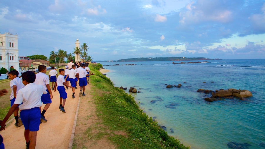 Camino costero con estudiantes uniformados, un faro y aguas rocosas.