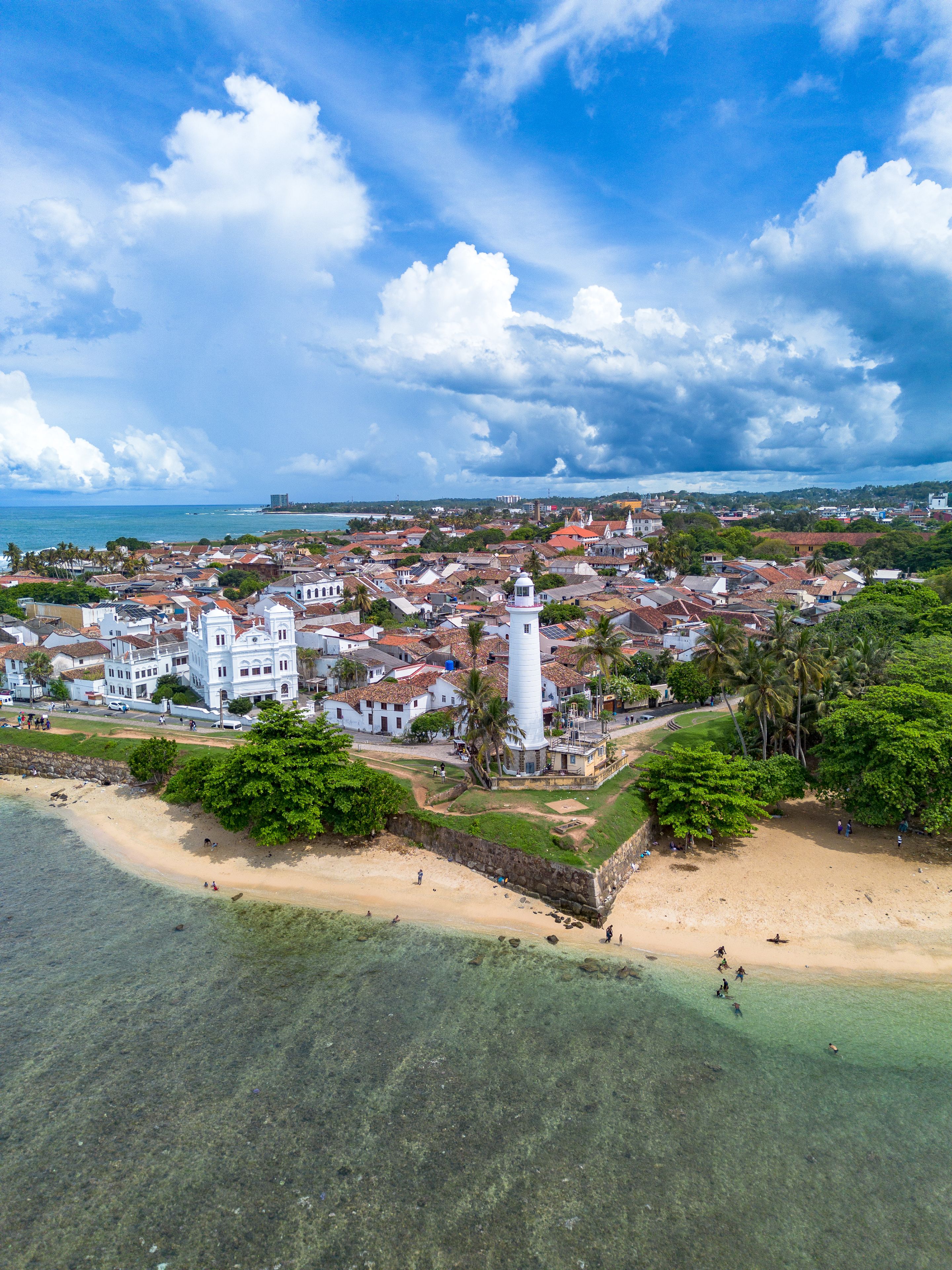 Vertical drone photo showing the tip of Galle Fort ramparts, Galle Lighthouse, pristine clear shallow coral waters, and the historic Dutch colonial townscape under a blue cloudy sky.