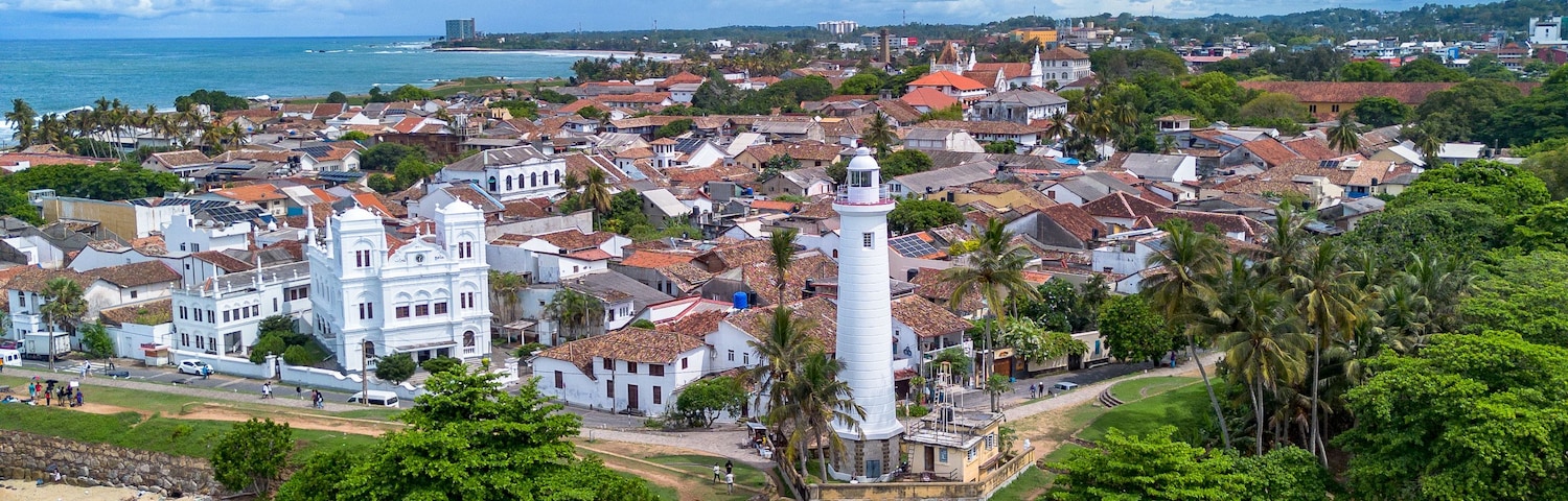 Vertical drone photo showing the tip of Galle Fort ramparts, Galle Lighthouse, pristine clear shallow coral waters, and the historic Dutch colonial townscape under a blue cloudy sky.