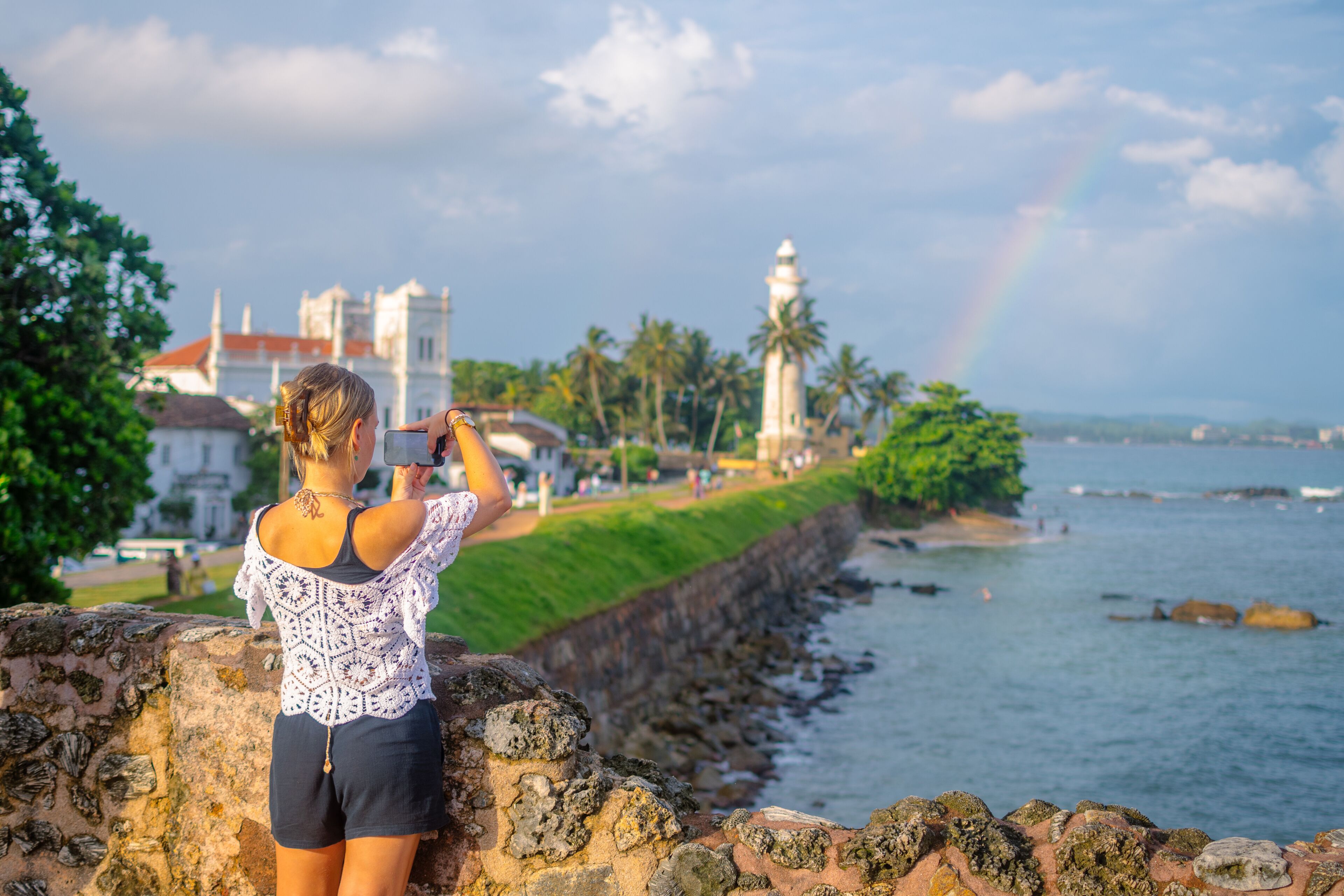 Female Tourist Taking Photo of Galle Fort Lighthouse Along the Coastal Promenade in Galle, Sri Lanka with Rainbow in the Sky