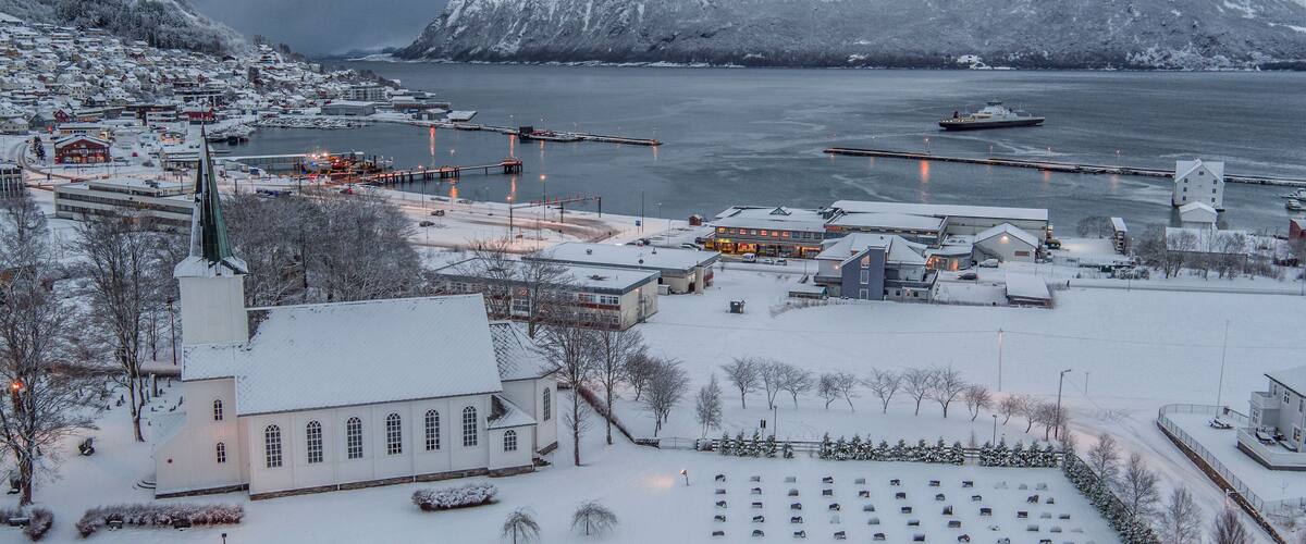 A snow-covered town by a fjord with a church, dock, and boats.