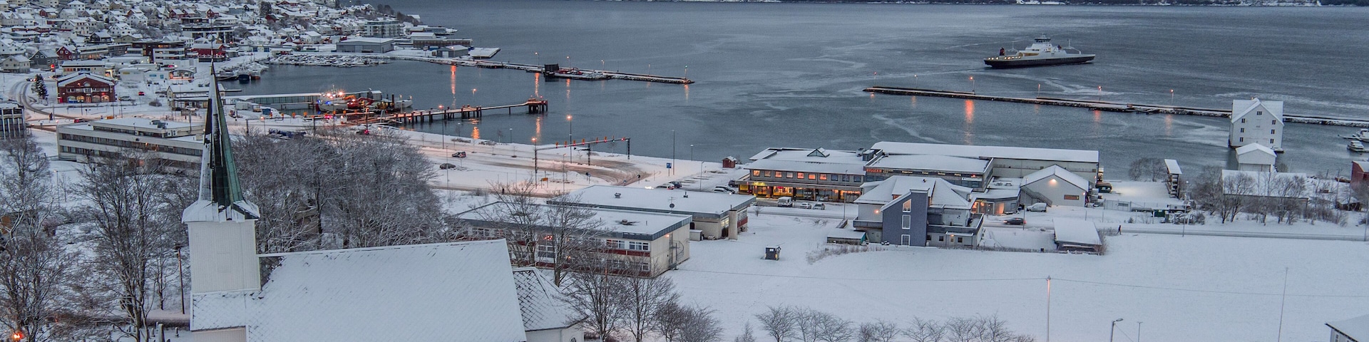 A snow-covered town by a fjord with a church, dock, and boats.