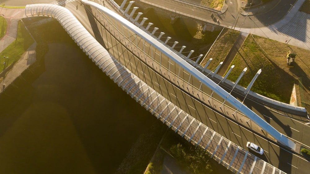 Un pont au design incurvé qui enjambe une étendue d’eau, entouré de verdure et d’infrastructures urbaines.
