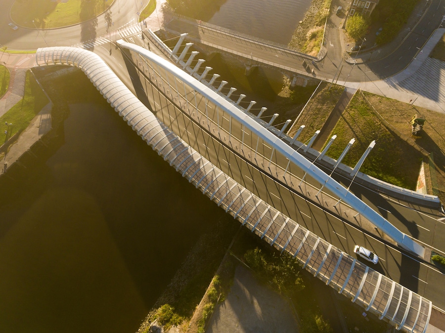 Een brug met een gebogen ontwerp over water, omgeven door groen en stedelijke infrastructuur.
