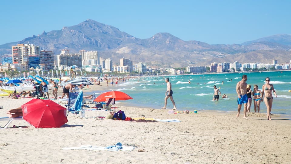 Una platja amb persones prenent el sol, ombrel·les i una muntanya al fons.