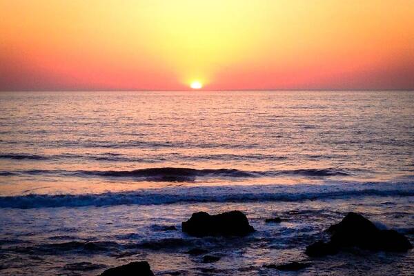 Ein Sonnenuntergang über dem Meer mit Wellen und Felsen im Vordergrund.