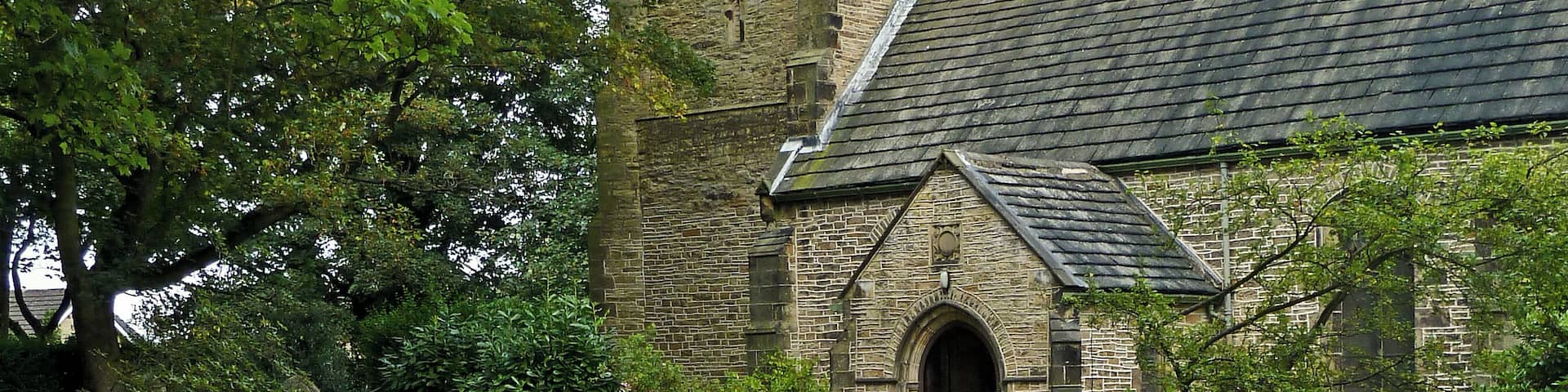 A church with a clock tower, a curved pathway lined with flowers, and a grassy area with trees and tombstones.