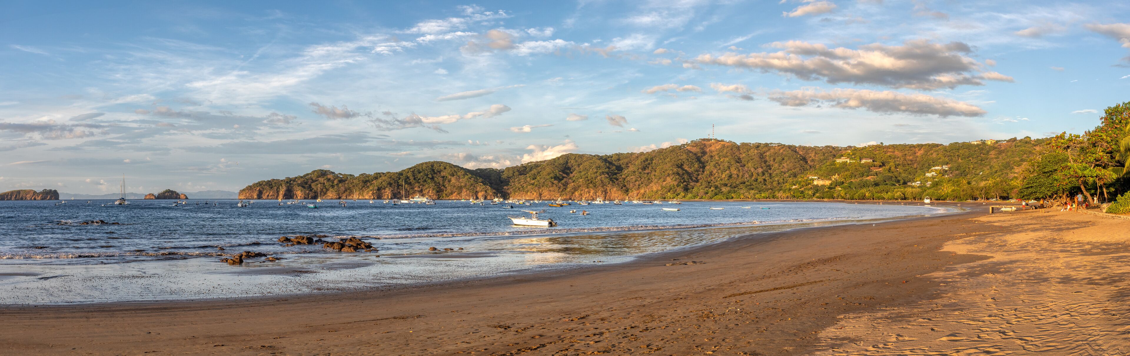 A beach with boats, a sandy shore, and a forested hill in the background.