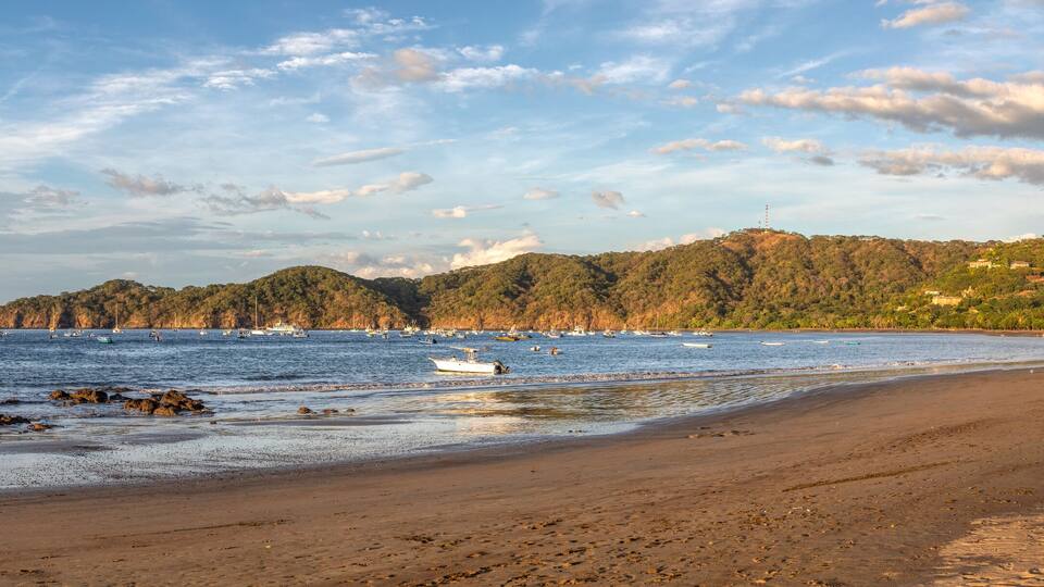 A beach with boats, a sandy shore, and a forested hill in the background.
