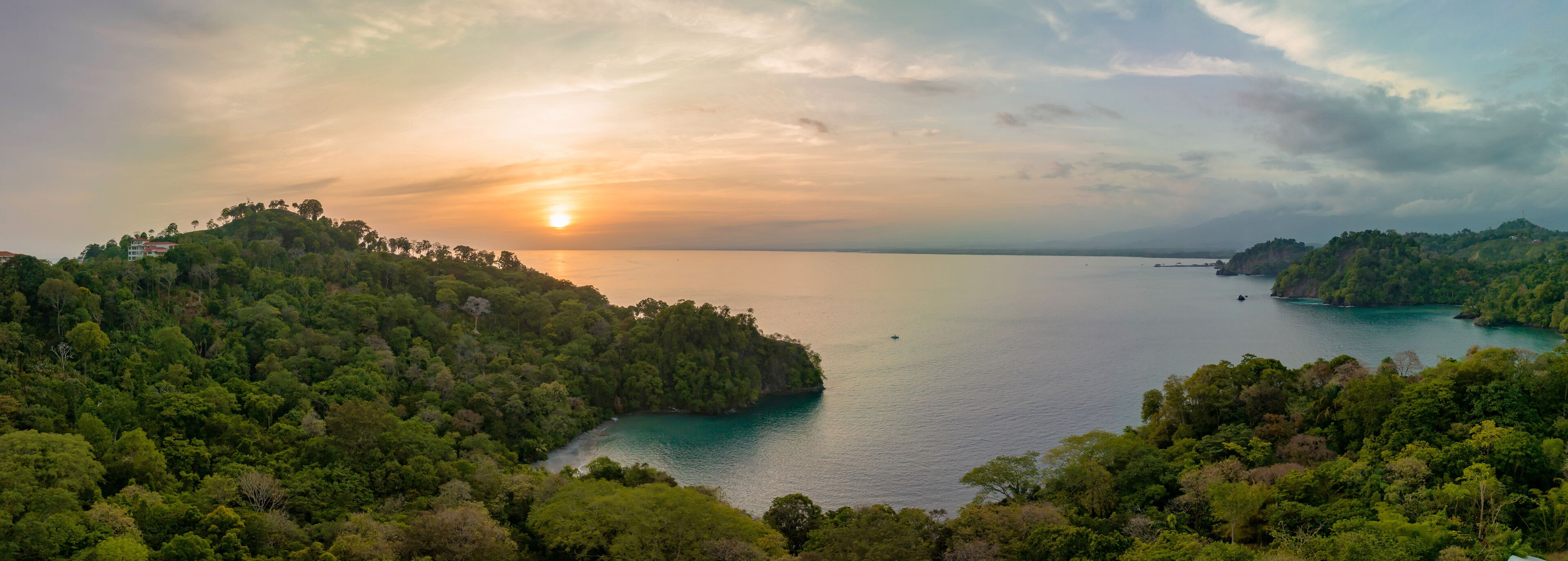 A coastal landscape with dense forested hills, a calm sea, and a setting sun.