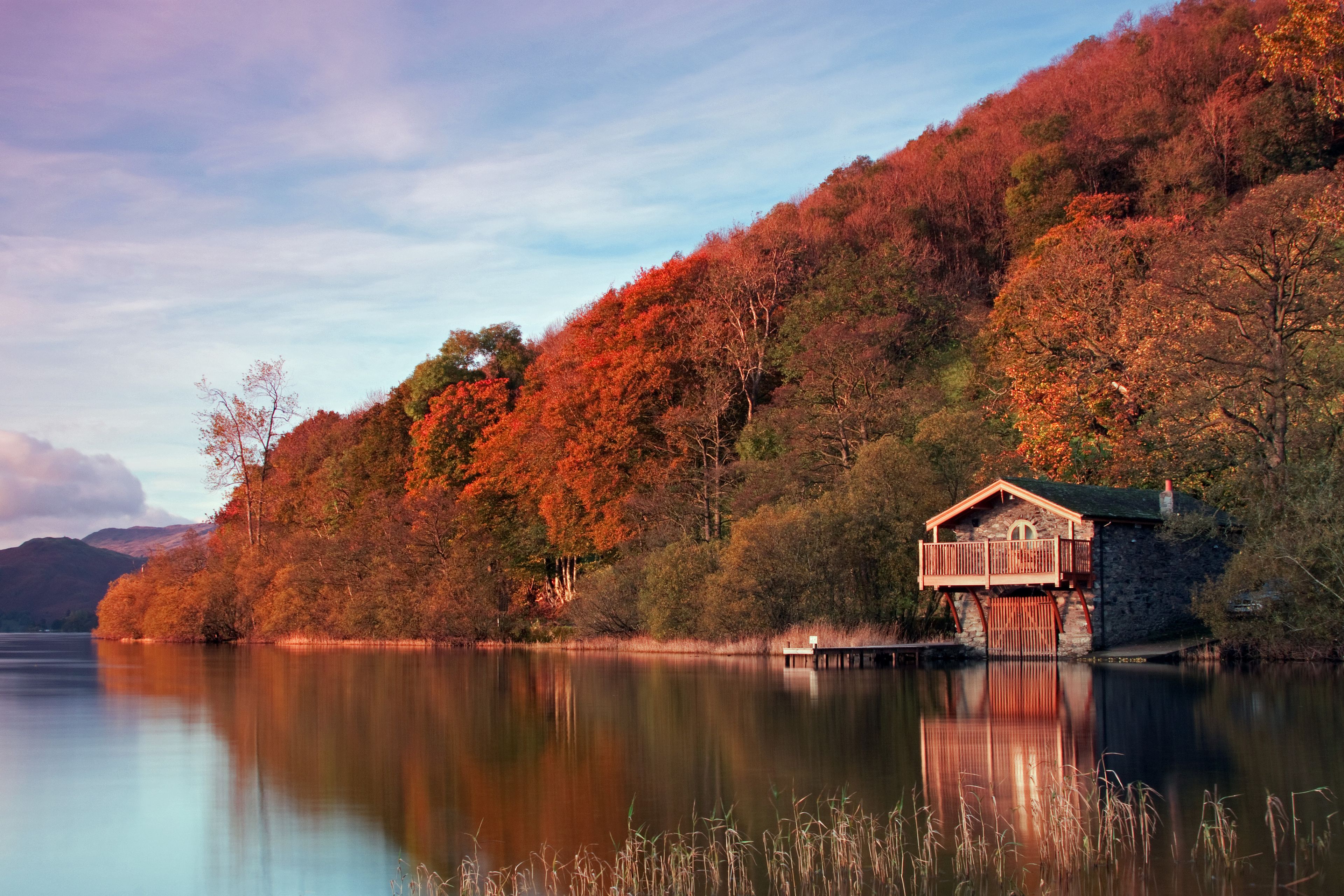 A lakeside cabin with a stone foundation and a wooden deck, surrounded by autumn-colored trees and a calm lake.