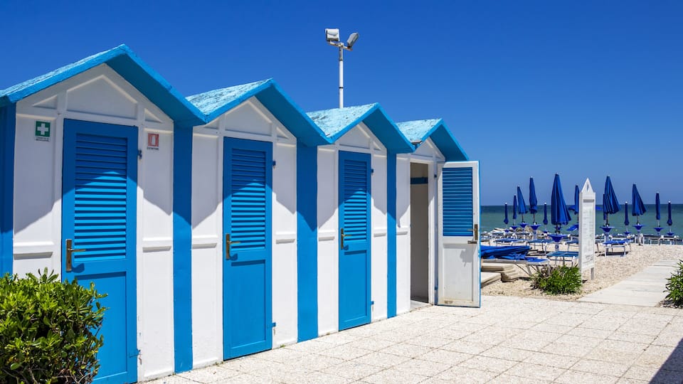 A row of blue and white beach huts with a clear blue sky and umbrellas in the background.