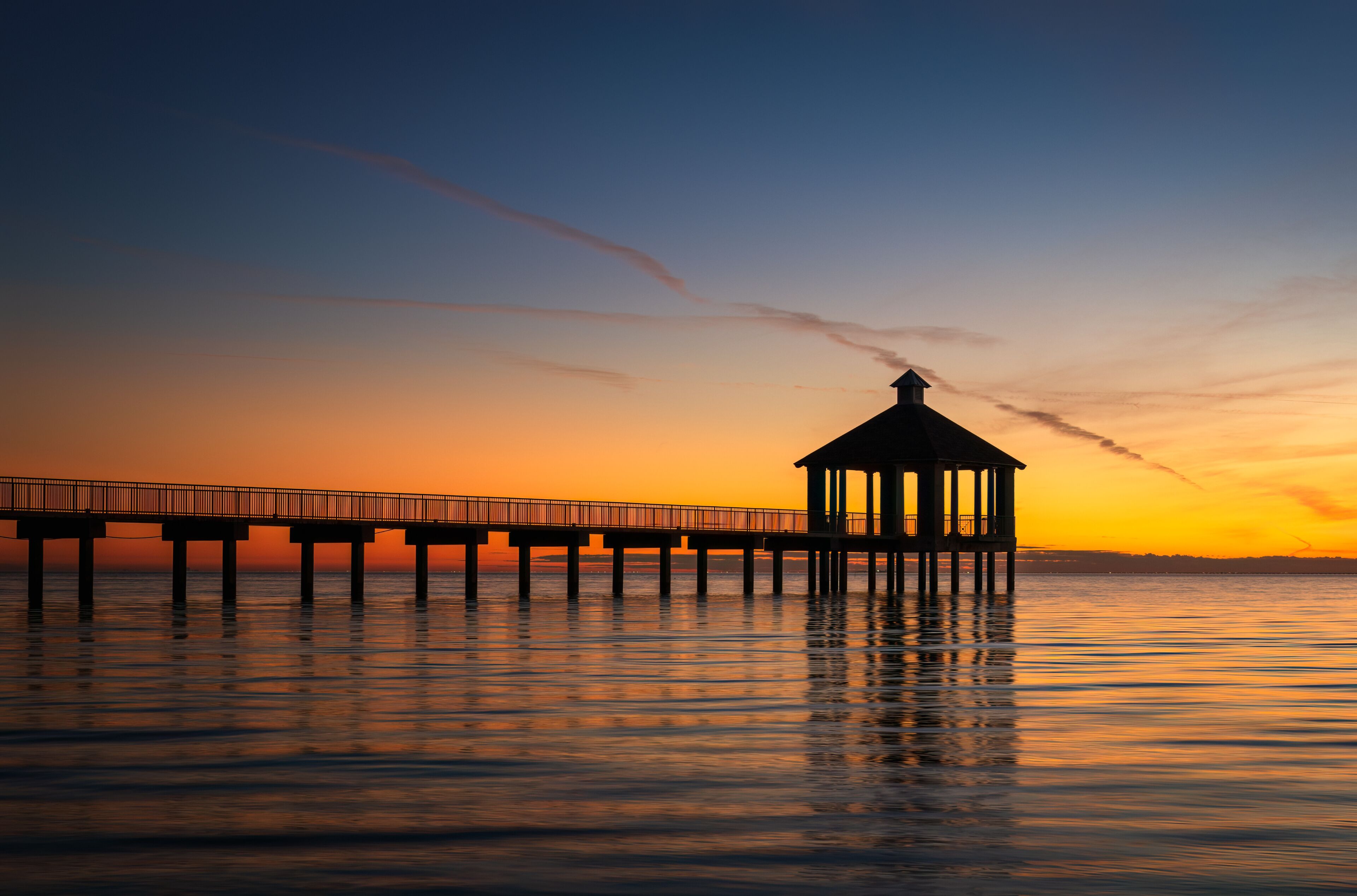 A pier extending into calm waters at sunset with a gazebo on the end.