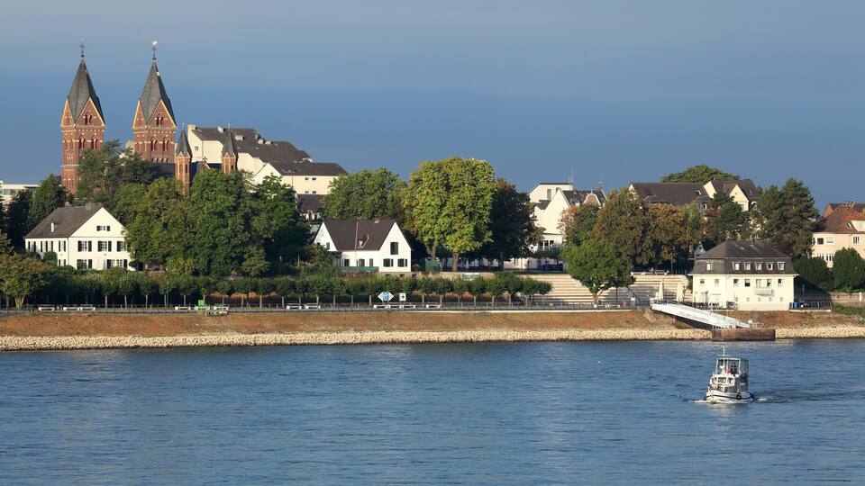 A river with a boat, a church with two towers, and residential buildings along the riverbank.