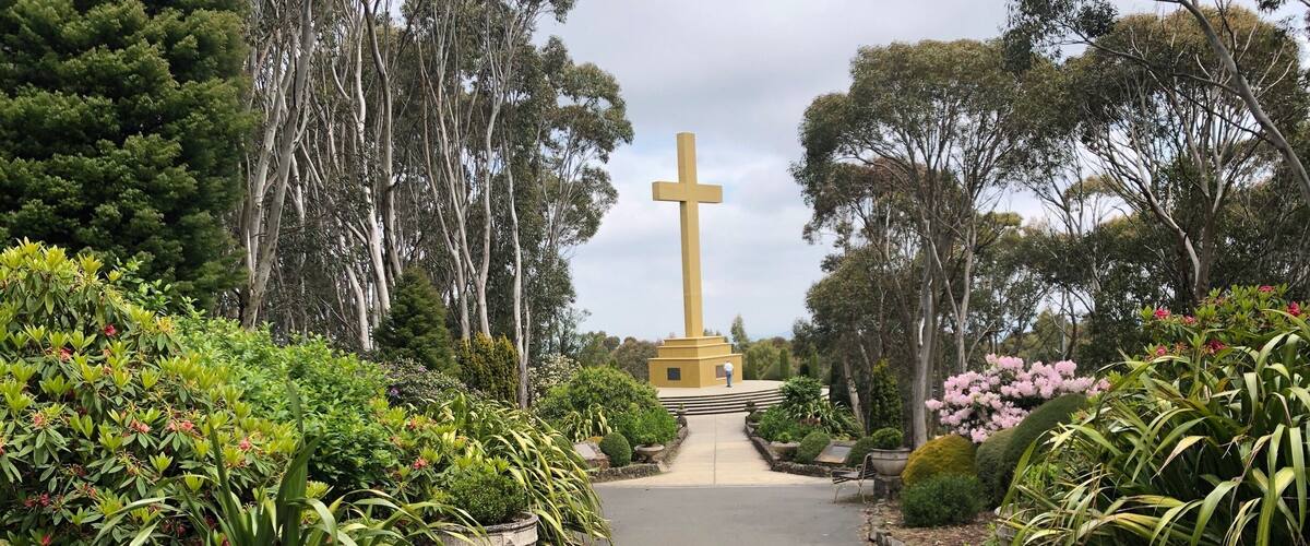 A pathway leading to a yellow monument with a cross, surrounded by lush greenery and flowering plants.