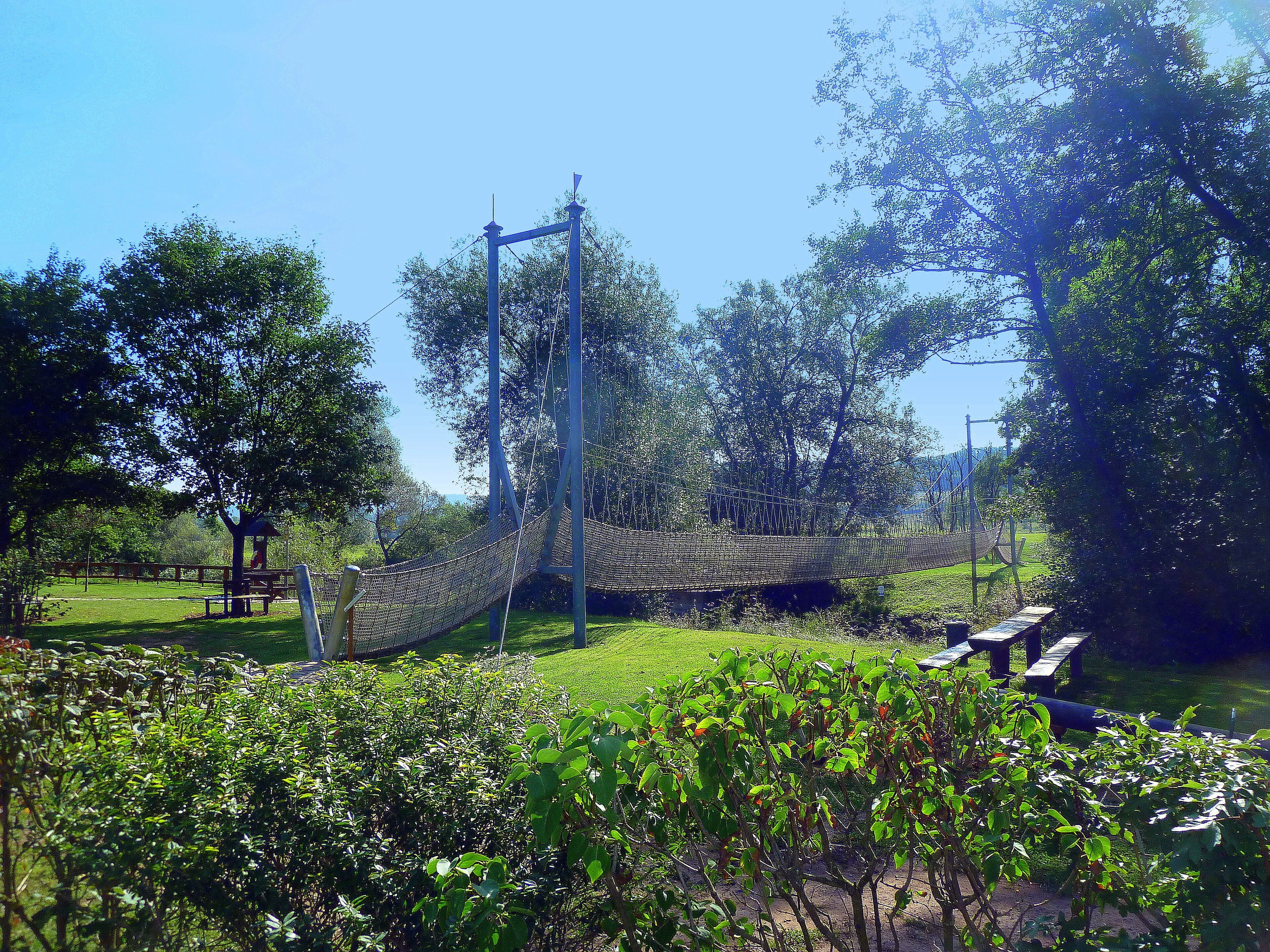 A park with a rope bridge, picnic tables, and lush greenery.