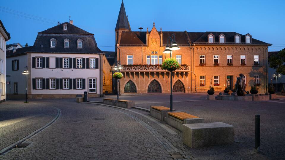 A cobblestone square with historic buildings, a church tower, and flower-adorned lampposts.