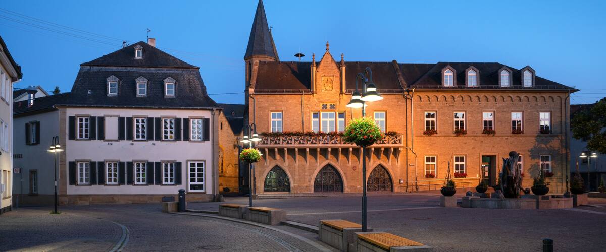 A cobblestone square with historic buildings, a church tower, and flower-adorned lampposts.