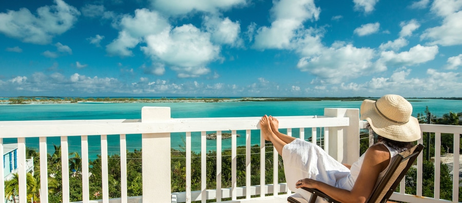Uma pessoa relaxando em uma varanda com vista para águas azul-turquesa e céu claro.