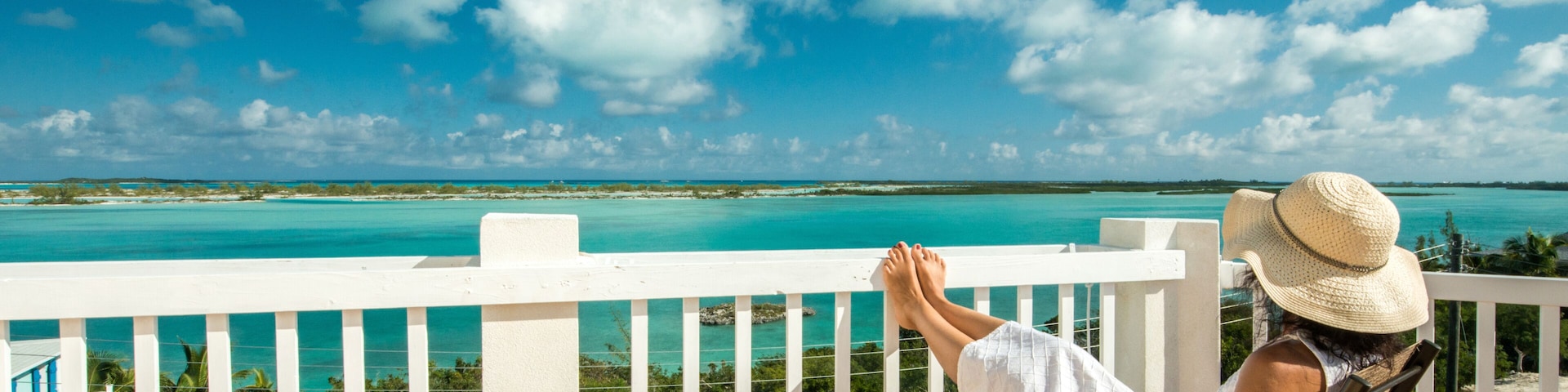 Uma pessoa relaxando em uma varanda com vista para águas azul-turquesa e céu claro.