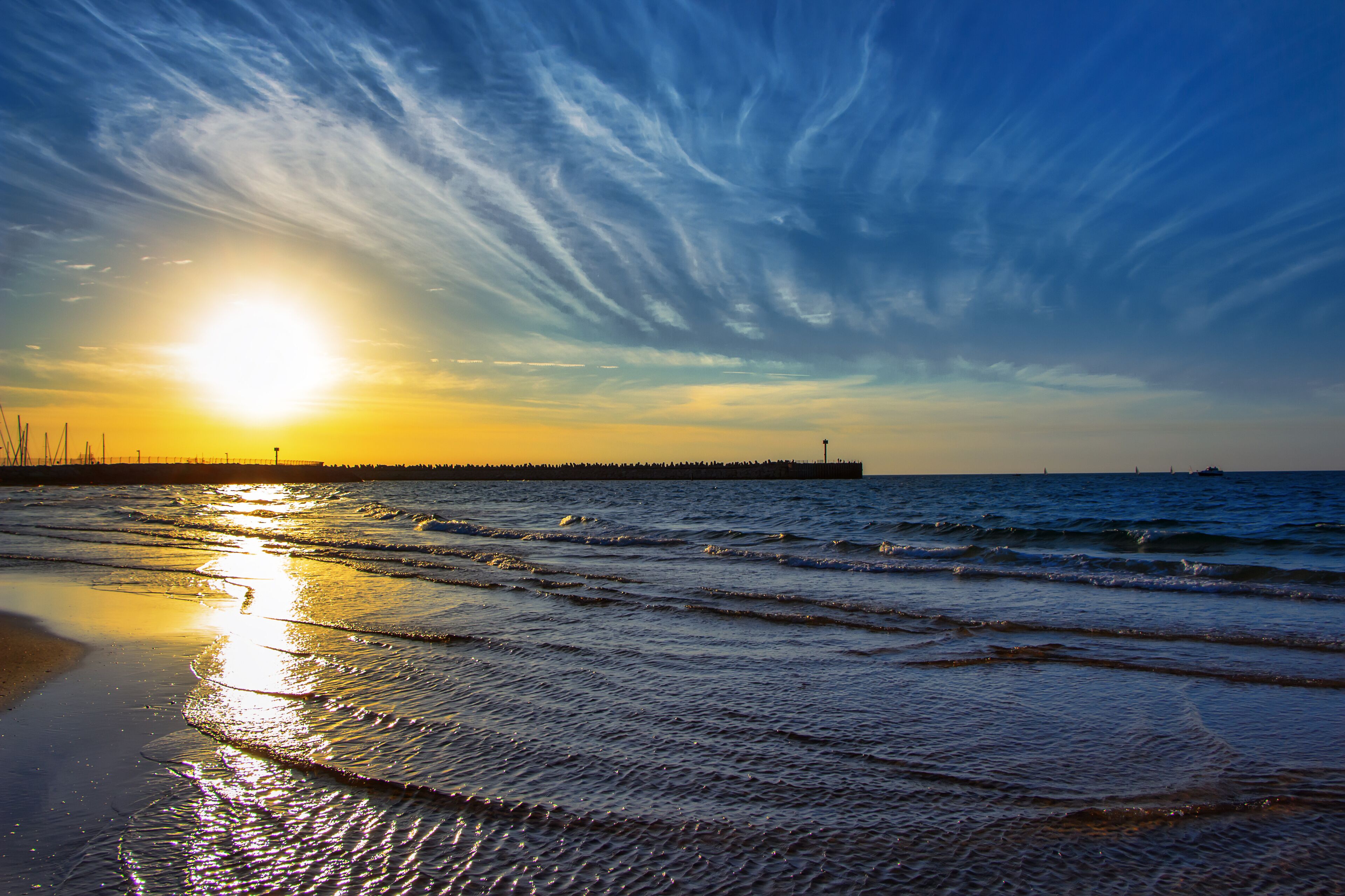 Uma praia com ondas, um pôr do sol e um cais ao longe.