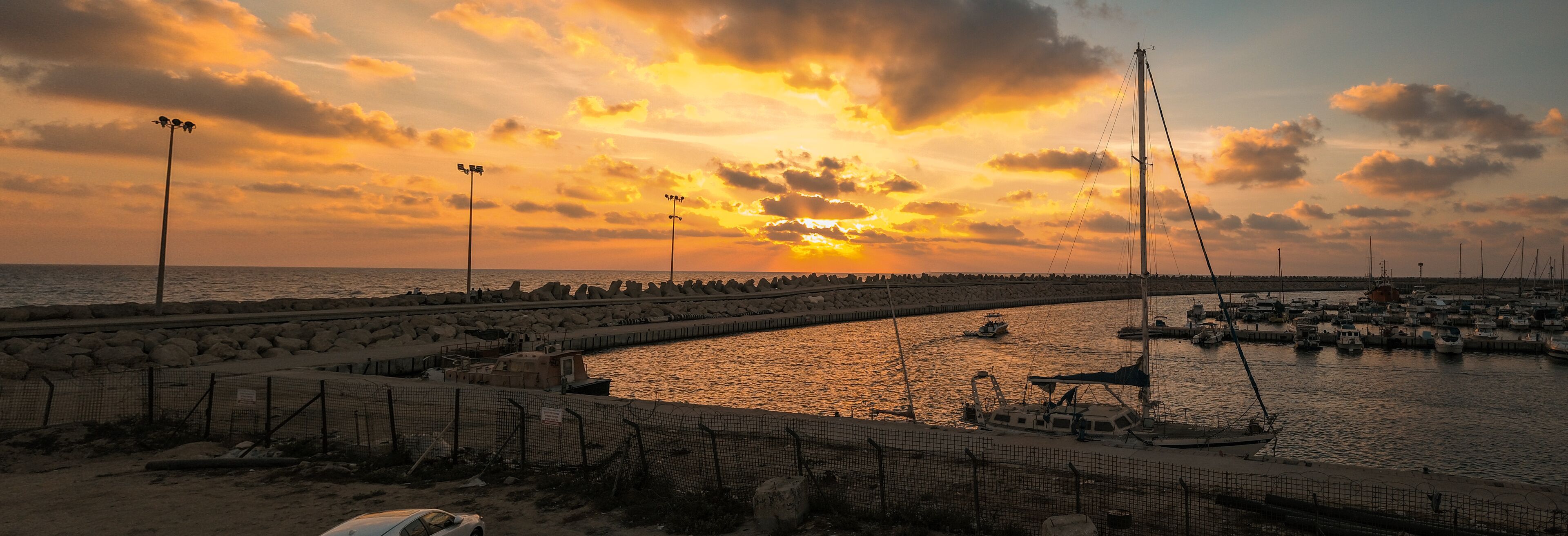 A sunset over a marina with boats and a rocky breakwater.