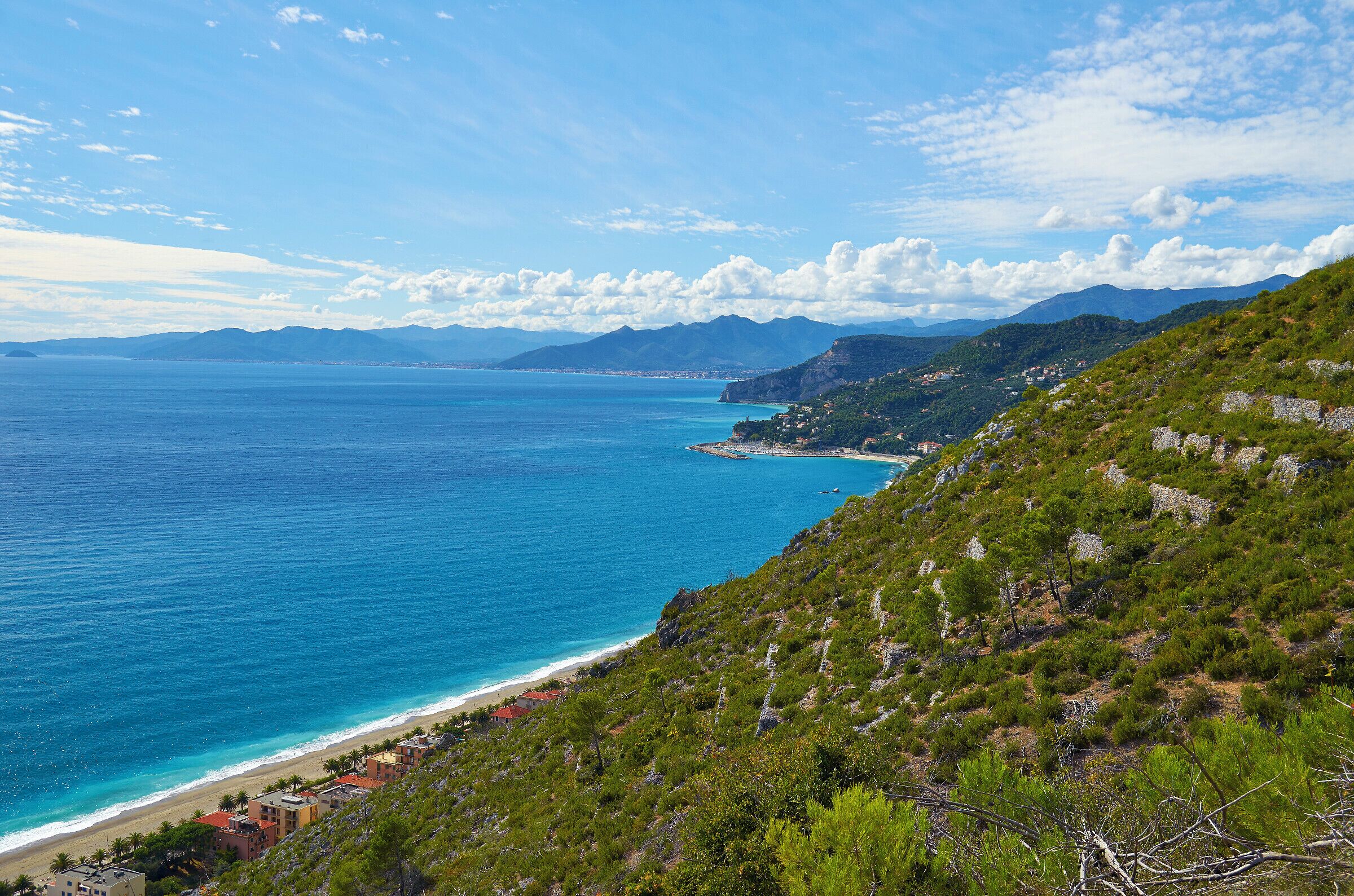 A coastal landscape with a clear blue sea, a sandy beach, and green hills.