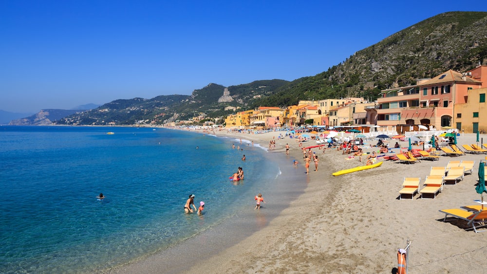 Ein sonniger Strand mit klarem, blauem Wasser, Menschen beim Schwimmen und Sonnenbaden sowie farbenfrohe Gebäude entlang der Küste.