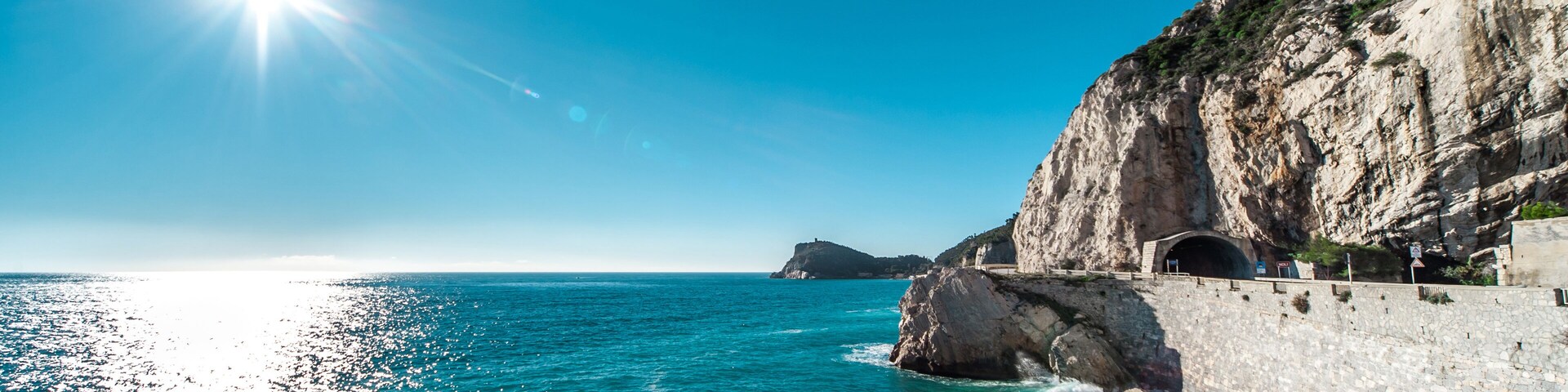 Escena costera con cielo azul despejado, un sol brillante y un acantilado rocoso con la entrada de un túnel.