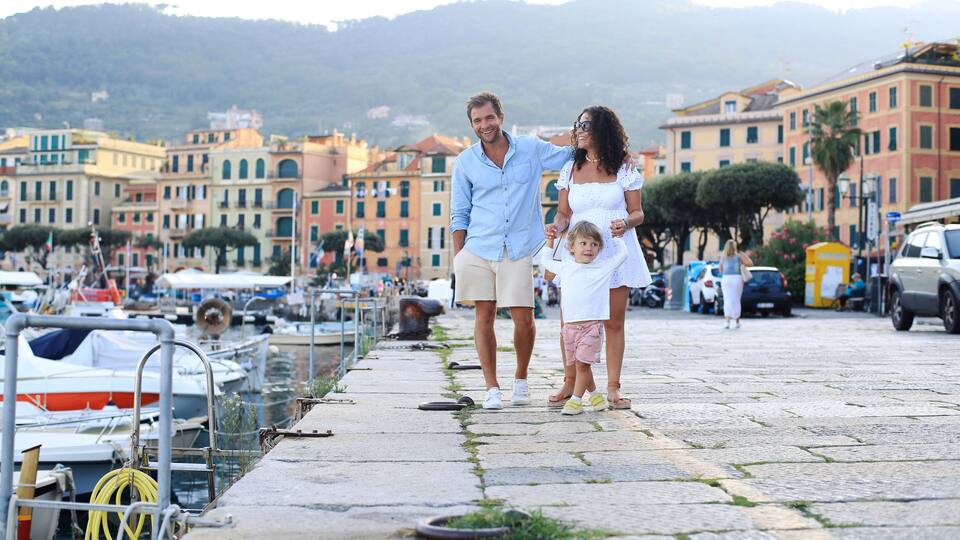 A waterfront promenade with boats, a stone pathway, and colorful buildings in the background.