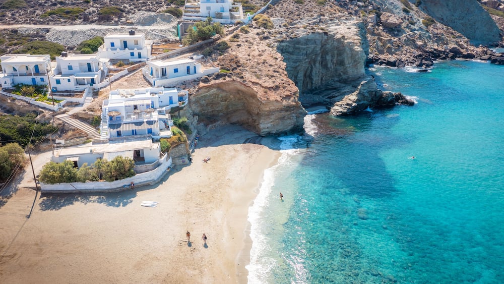 A coastal village with white buildings, a sandy beach, and clear blue waters.