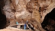 Una grotta con stalattiti e stalagmiti, una persona seduta su una sporgenza e un'altra in piedi su una piattaforma.