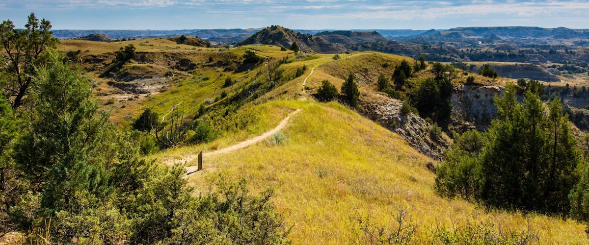 A mountainous landscape with a dirt path, green vegetation, and distant hills under a blue sky with clouds.