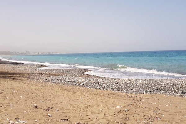 Μια παραλία με βότσαλα, κύματα, καθαρό ουρανό και κτίρια που διακρίνονται στο βάθος.