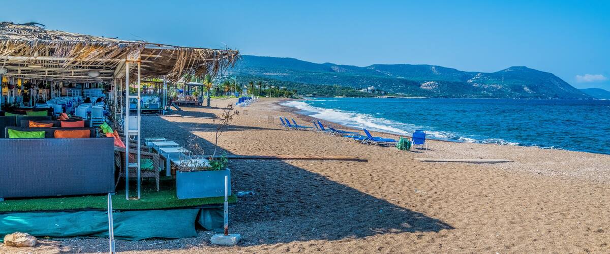 A beach with a thatched-roof structure, blue lounge chairs, and a mountainous backdrop.