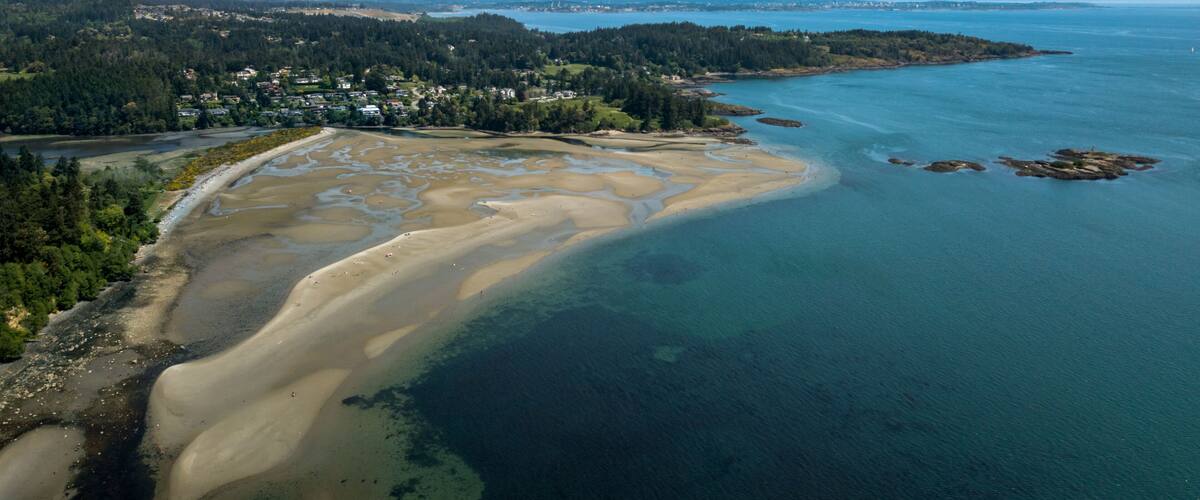 An aerial view of a coastal area with a sandy beach, clear blue waters, and a forested peninsula.