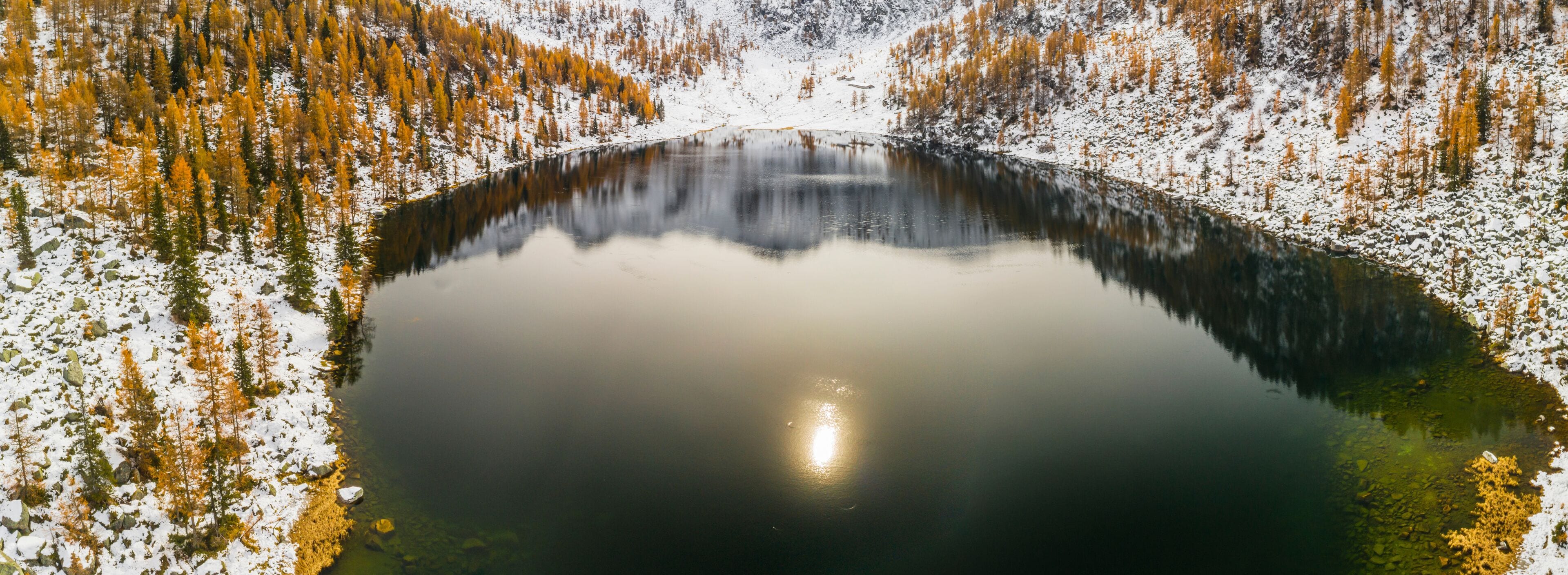 Aerial view of San Giuliano  lake reflecting mountains, surrounded by snow-dusted golden trees. San Giuliano Lakes,Adamello Brenta Natural Park,Caderzone Terme,Trentino,Italy